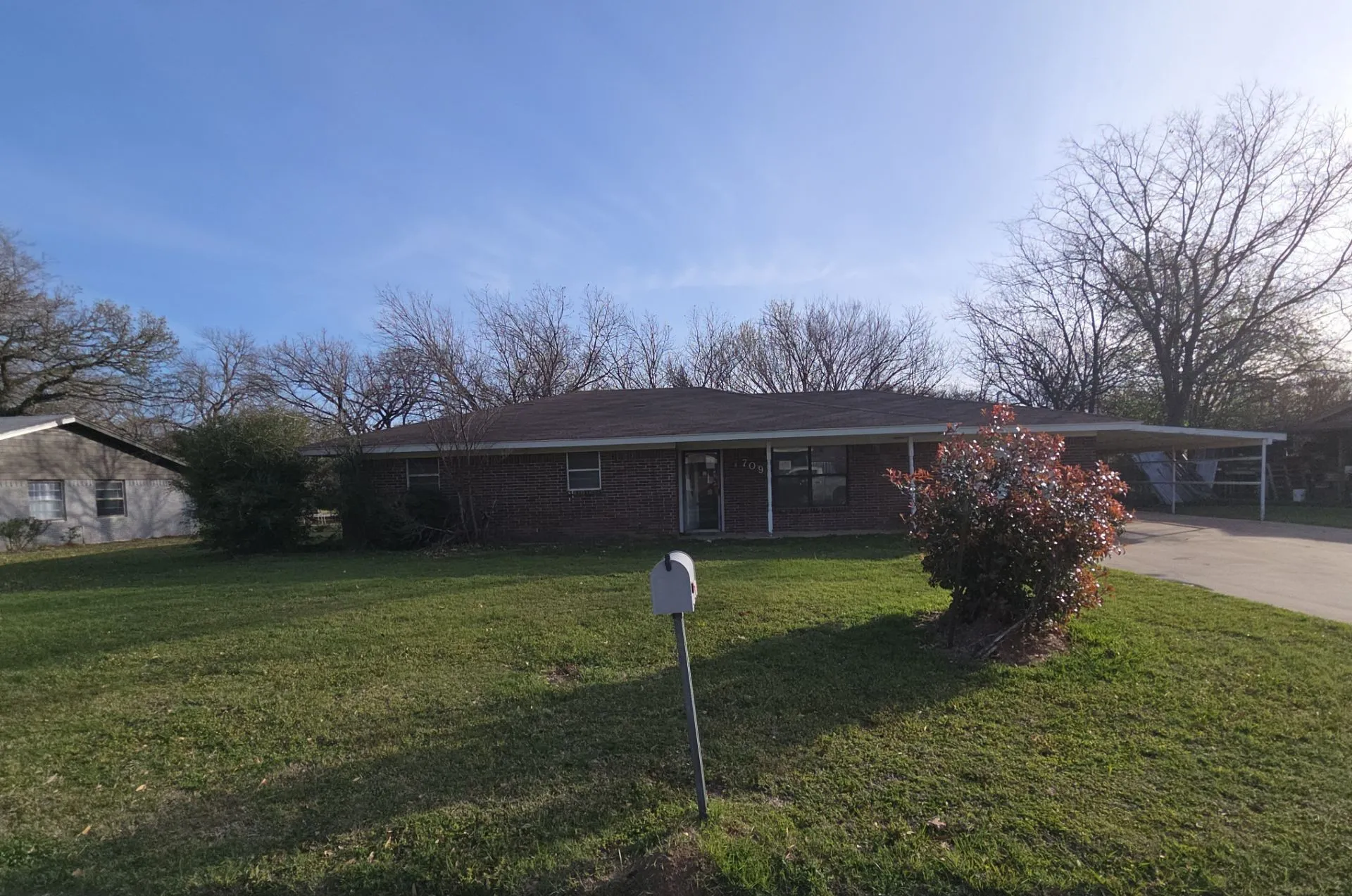 Brick exterior home featuring a covered carport, mature landscaping, and a broad front lawn