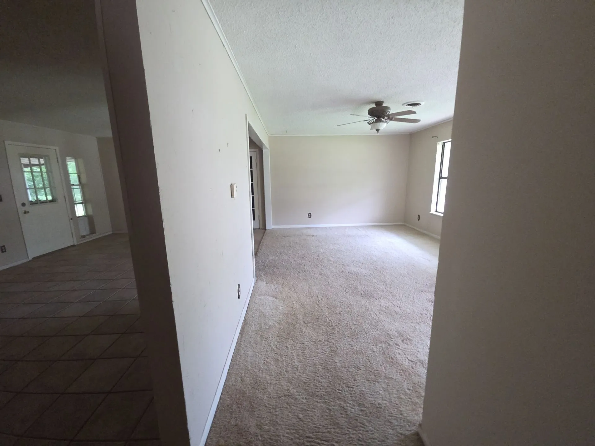 Spacious interior room featuring light-colored wall paint, textured ceiling, light beige carpeting, and a ceiling fan with integrated lighting