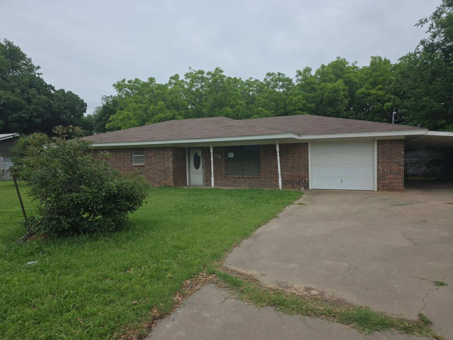 Brick exterior home featuring an attached garage, a carport, and a front yard with mature landscaping