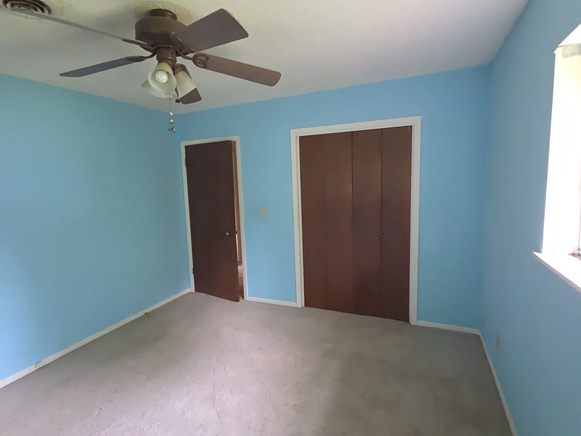 Room with light blue walls, light-colored carpet, a ceiling fan with integrated lighting, and a window with white trim
