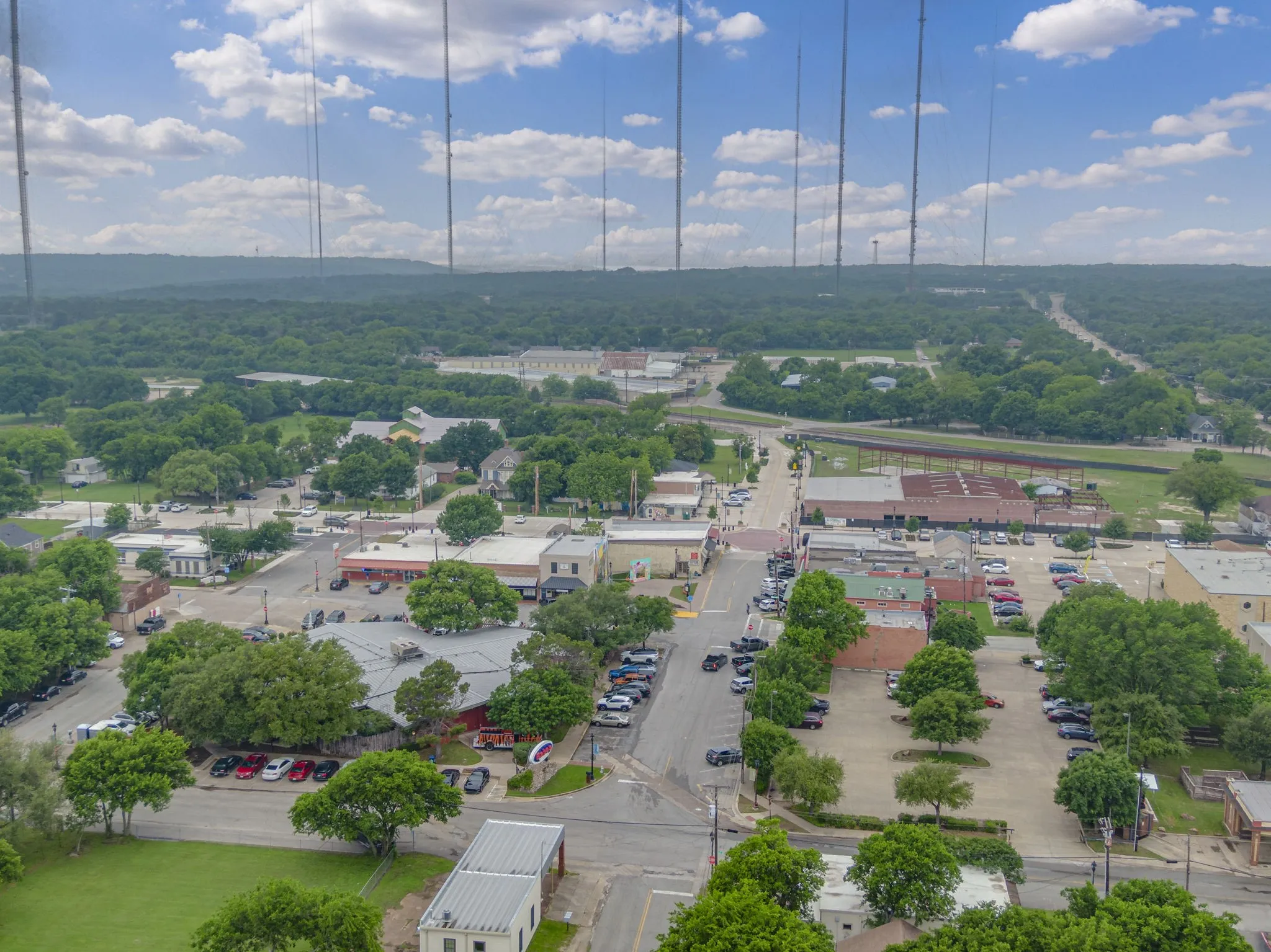 Aerial perspective showcasing a vibrant commercial district with surface parking, surrounding green spaces, and a distant tree-lined horizon