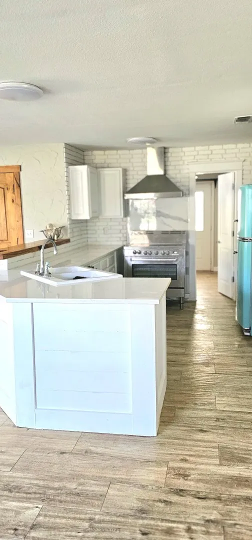 Kitchen featuring an island with an integrated sink, white cabinetry, wood-finish flooring, a stainless steel range, and a decorative tile backsplash