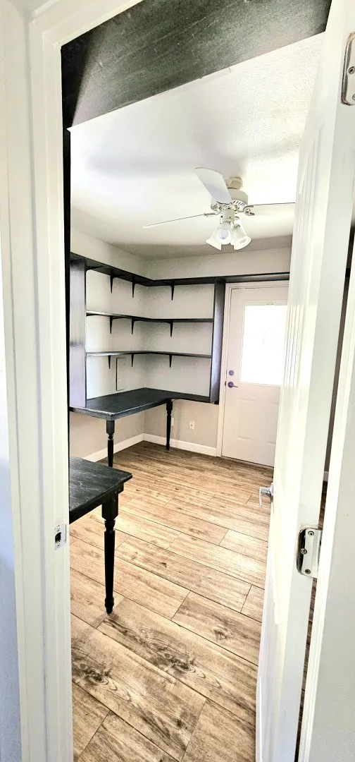 Versatile interior room featuring wood-finish flooring, built-in shelving, integrated desk space, a ceiling fan with light fixture, and a white exterior door