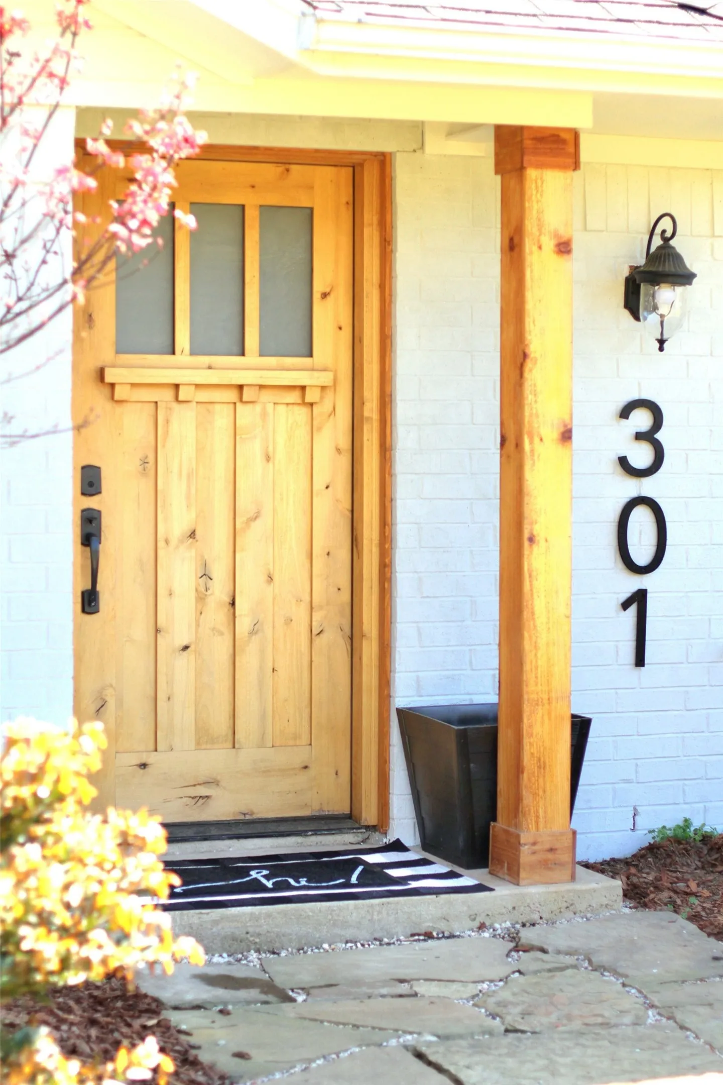 Natural wood entry door with upper window panes, complemented by a substantial wood support post and white brick exterior