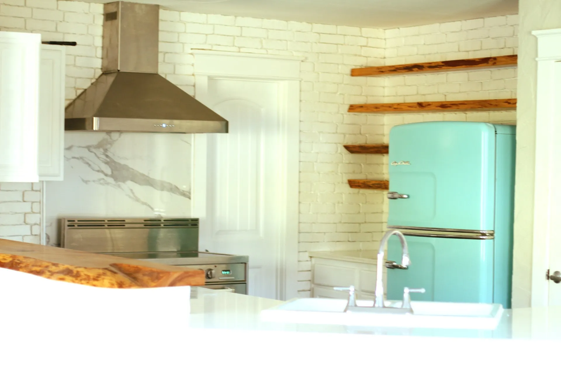 Kitchen featuring a stainless steel range hood, white brick-patterned walls, a white stone-look backsplash, white cabinetry, and open wood shelving