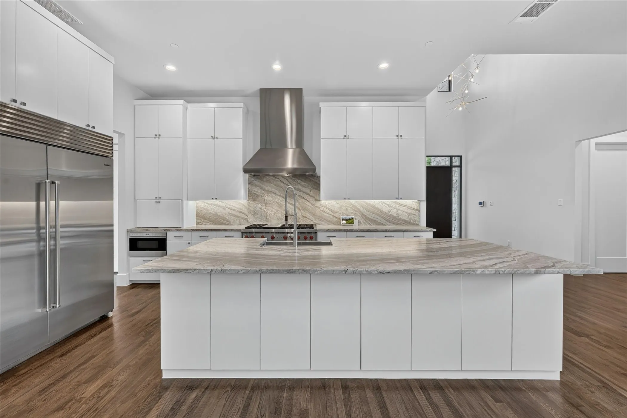 Modern kitchen featuring a large central island with a natural stone countertop, white cabinetry, integrated stainless steel appliances, and wood-finish flooring