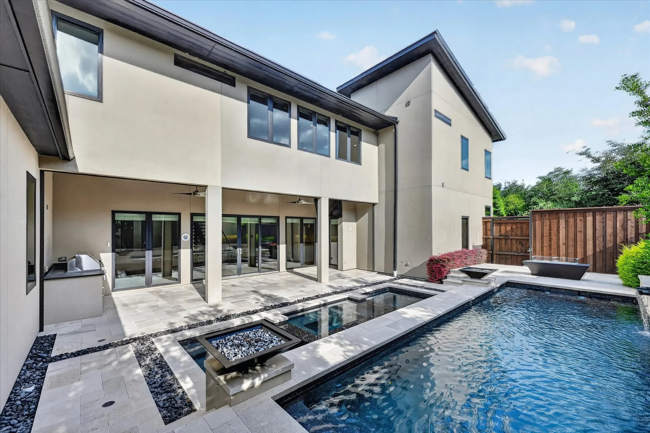 Rear exterior featuring a spacious covered patio, a multi-level pool with integrated spa, a built-in outdoor grill, and a fire pit feature