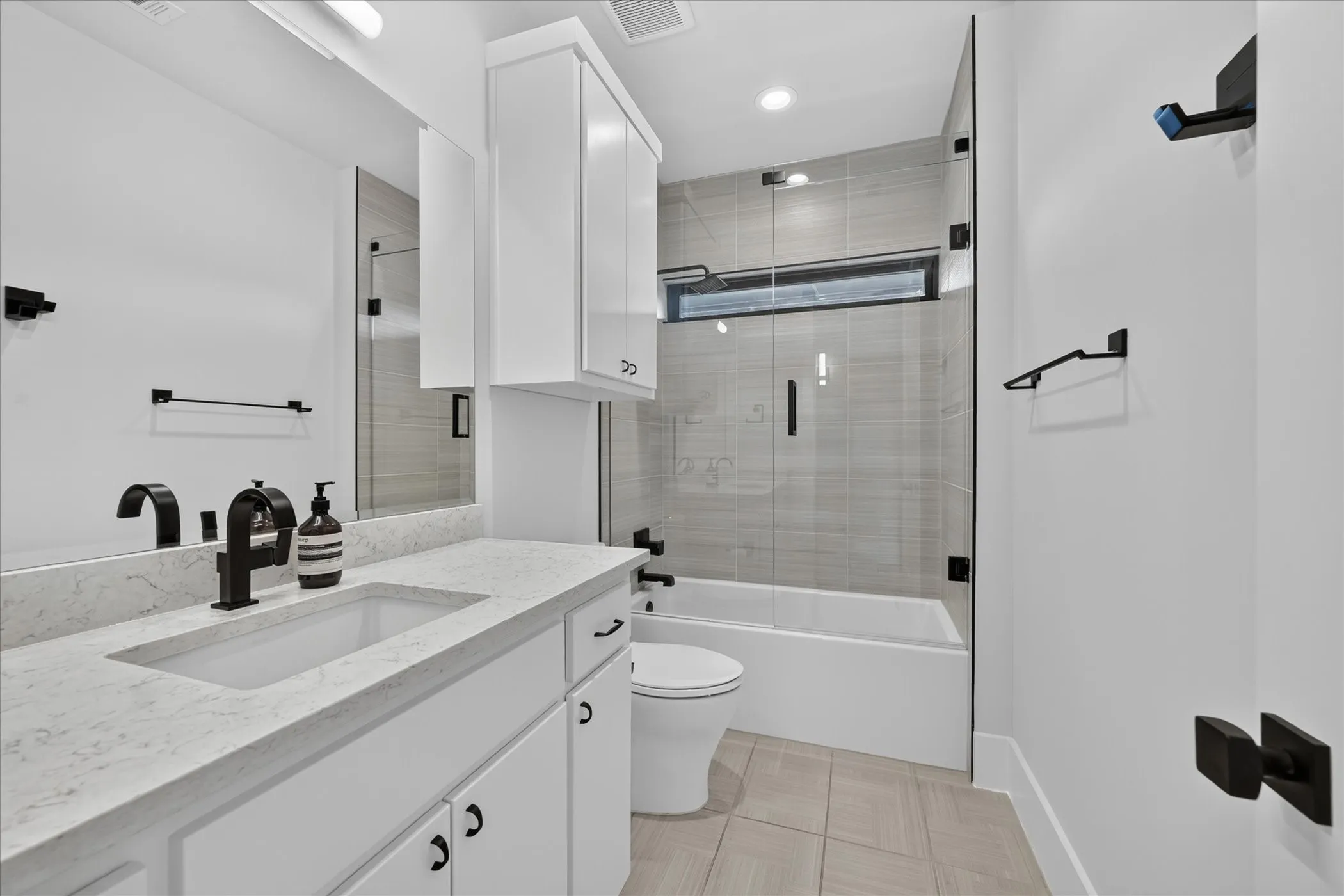 Bathroom featuring a quartz-finish countertop vanity with an integrated sink, matte black fixtures, white cabinetry, and a glass-enclosed shower-tub combination with tile surround