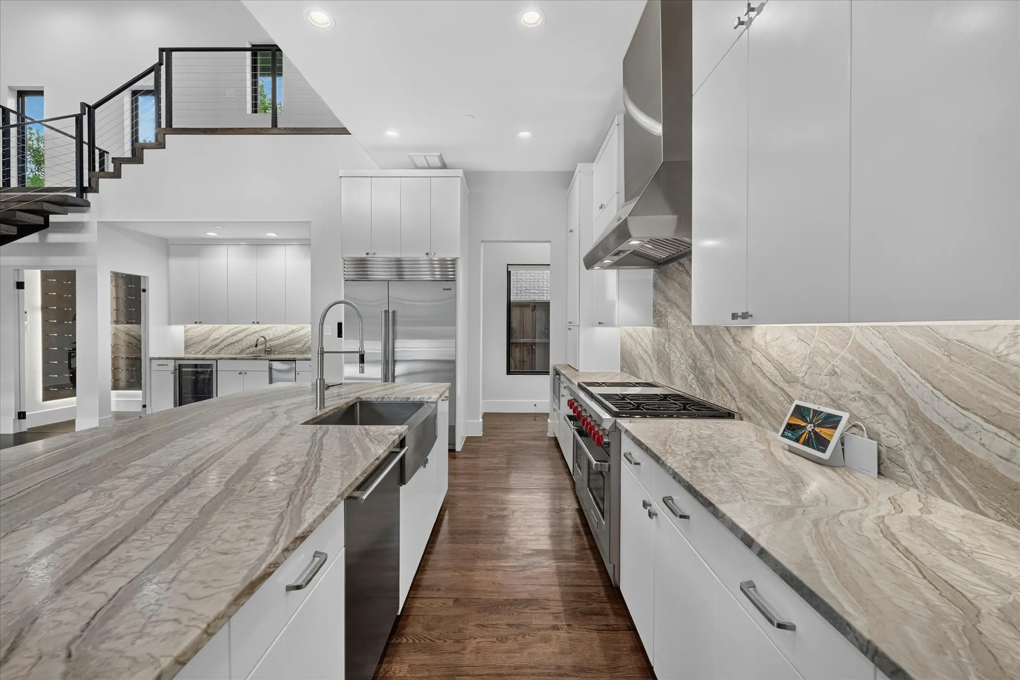 Open-concept kitchen featuring an oversized island with a farmhouse sink, integrated stainless steel appliances, premium stone countertops and backsplash, and rich wood-finish flooring