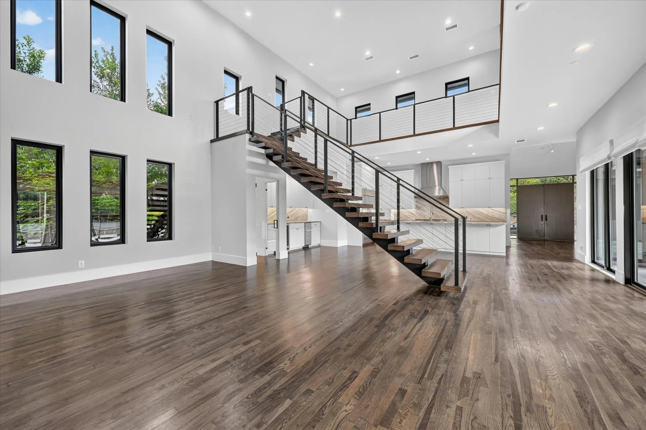Open-concept living space featuring wood-finish flooring, a contemporary floating staircase with metal railings, a kitchen area with light cabinetry and stone countertops, and multiple windows with dark frames