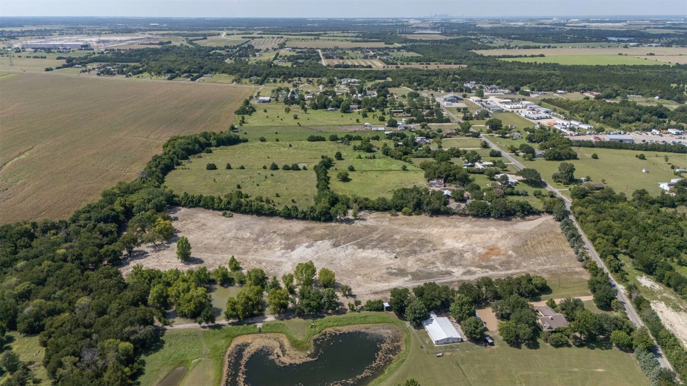 Expansive land parcel featuring a cleared building site, mature trees, a natural pond, and surrounding open fields
