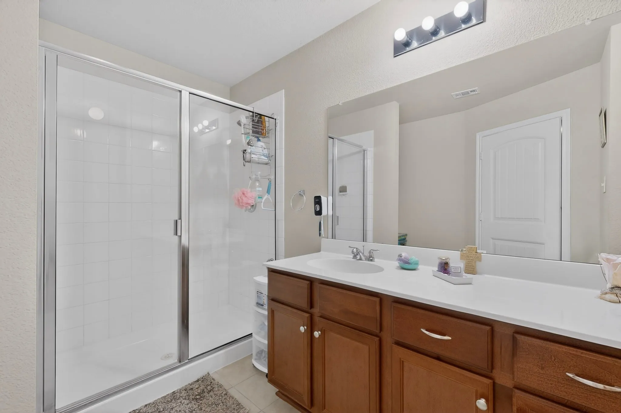 Bathroom featuring a glass-enclosed shower with white tiled walls, a single vanity with a white countertop and integrated sink, a large wall-mounted mirror, and wood-finish cabinetry with silver-tone hardware