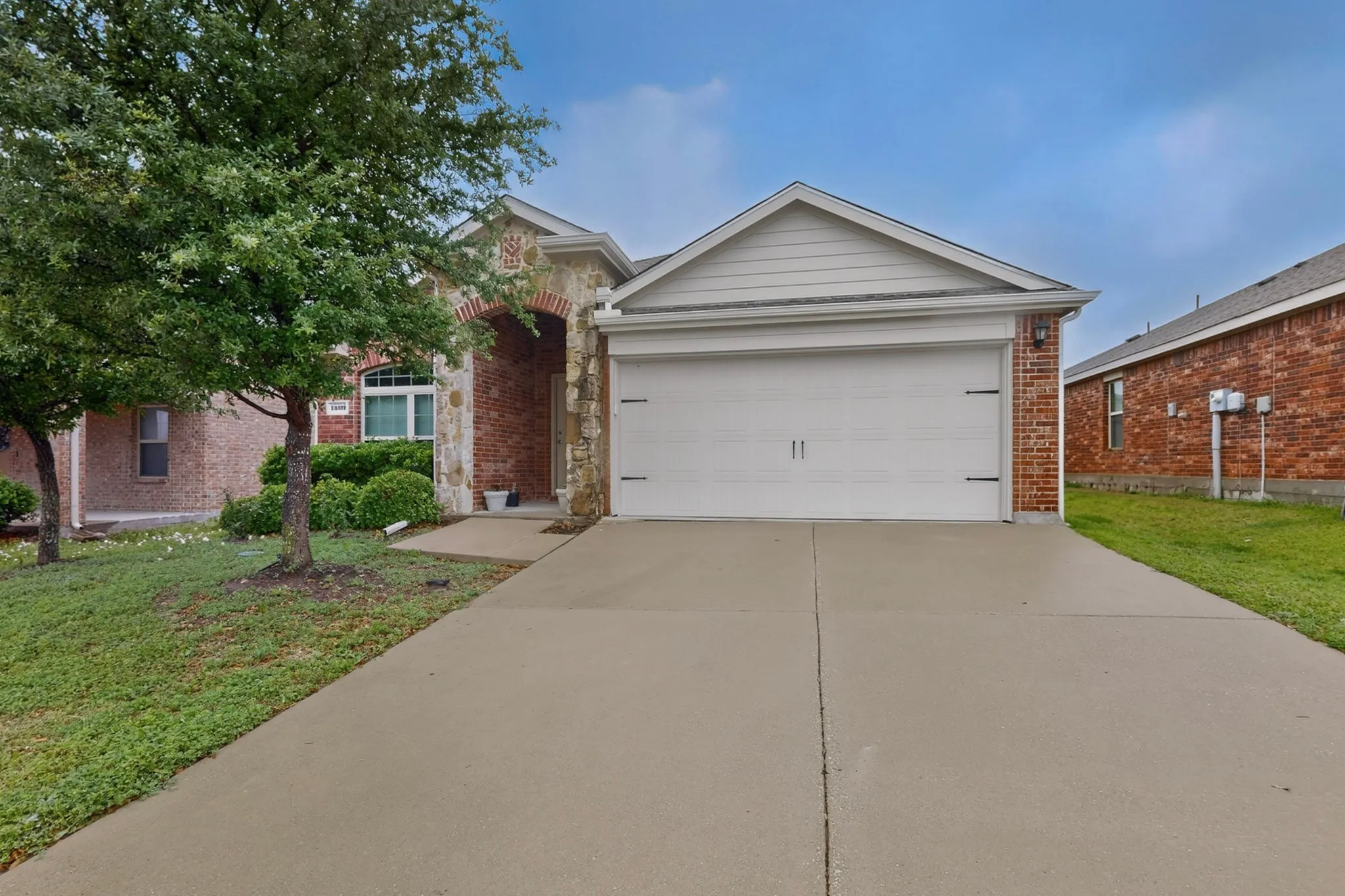 Brick and stone facade featuring an arched entry, an attached garage with a white door, a concrete driveway, and established landscaping
