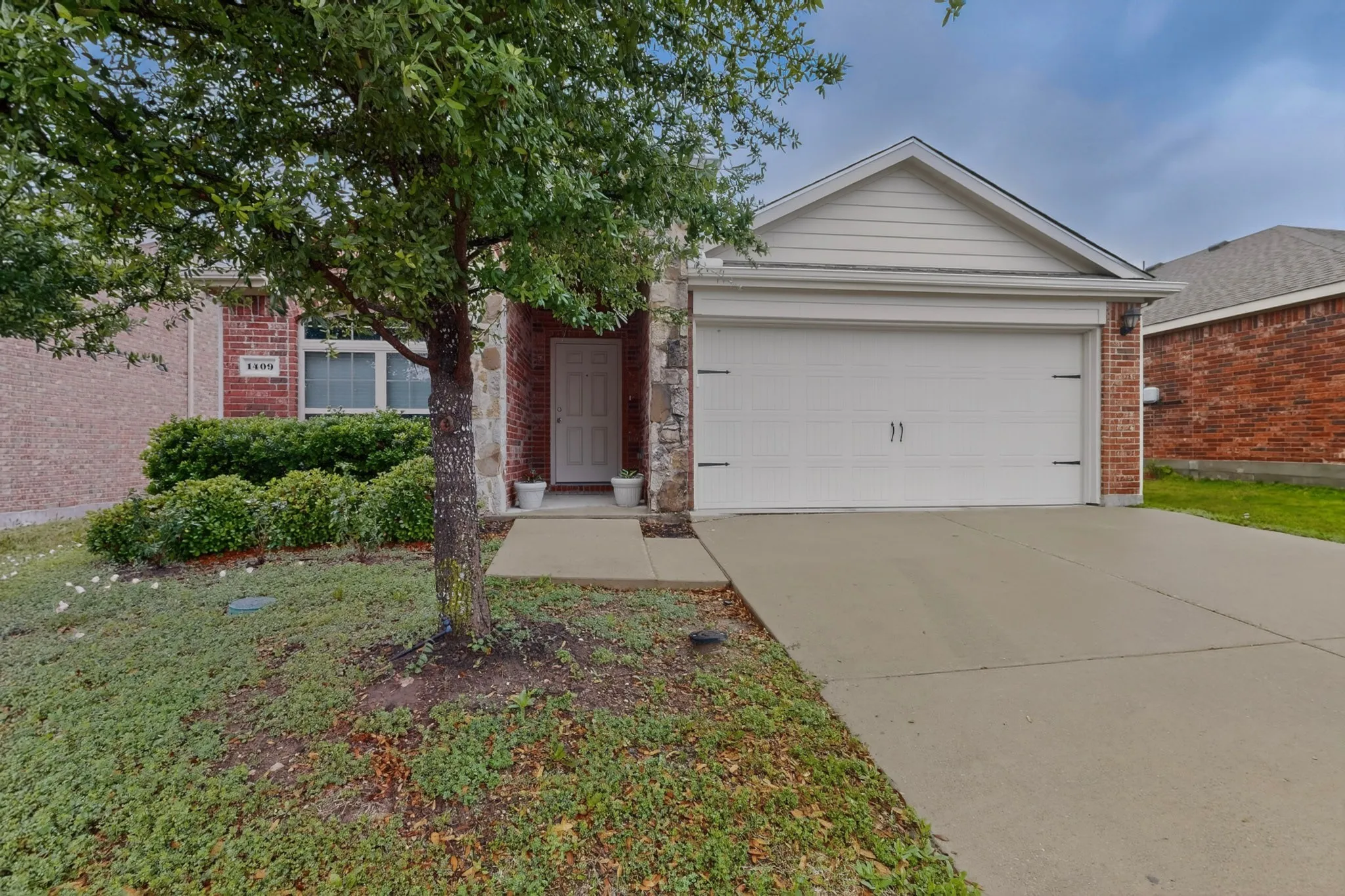 Brick and stone facade featuring a two-car garage, mature front yard tree, concrete driveway, and front entry with white door