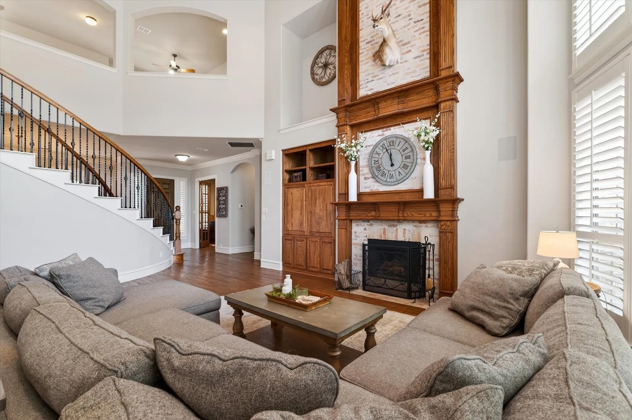 The floor-to-ceiling brick fireplace with wood mantle anchors the room, while the open staircase and tall windows add architectural drama.