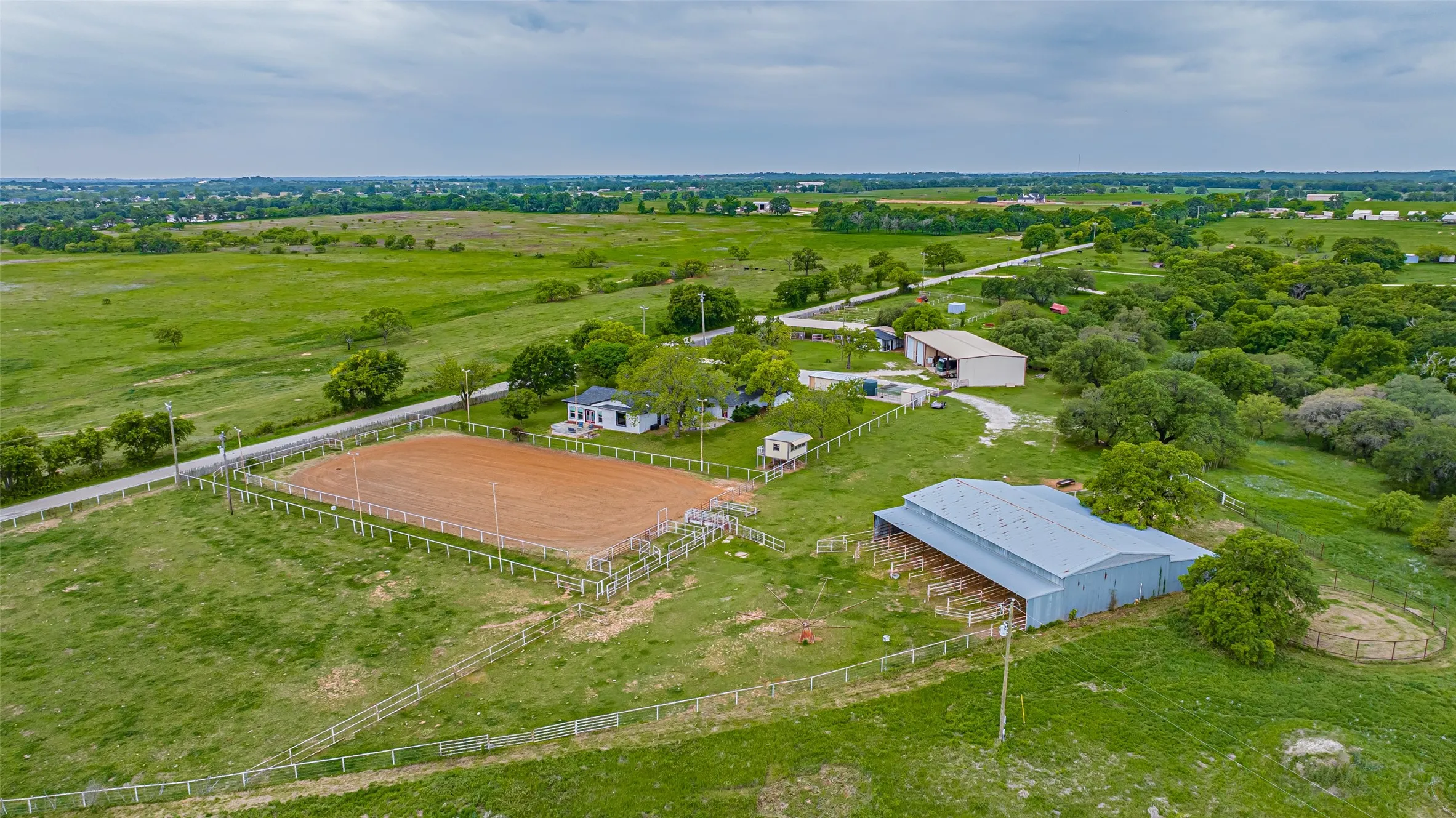 Expansive property featuring a fenced riding arena, a primary residence, a large barn structure with stalls, an additional outbuilding, and mature trees across a verdant landscape