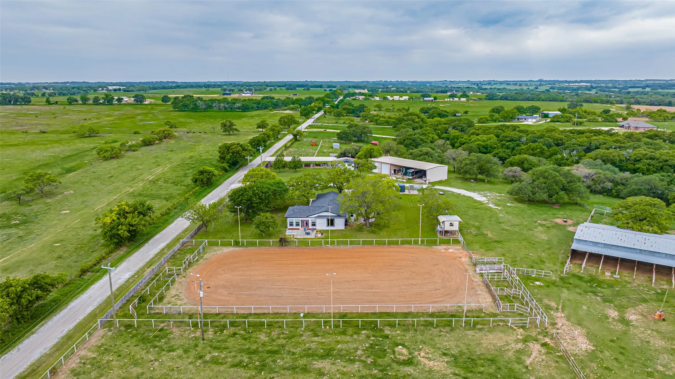 Expansive outdoor equestrian arena with perimeter fencing, a white single-story residence with a dark roof, a large metal barn, and a substantial outbuilding