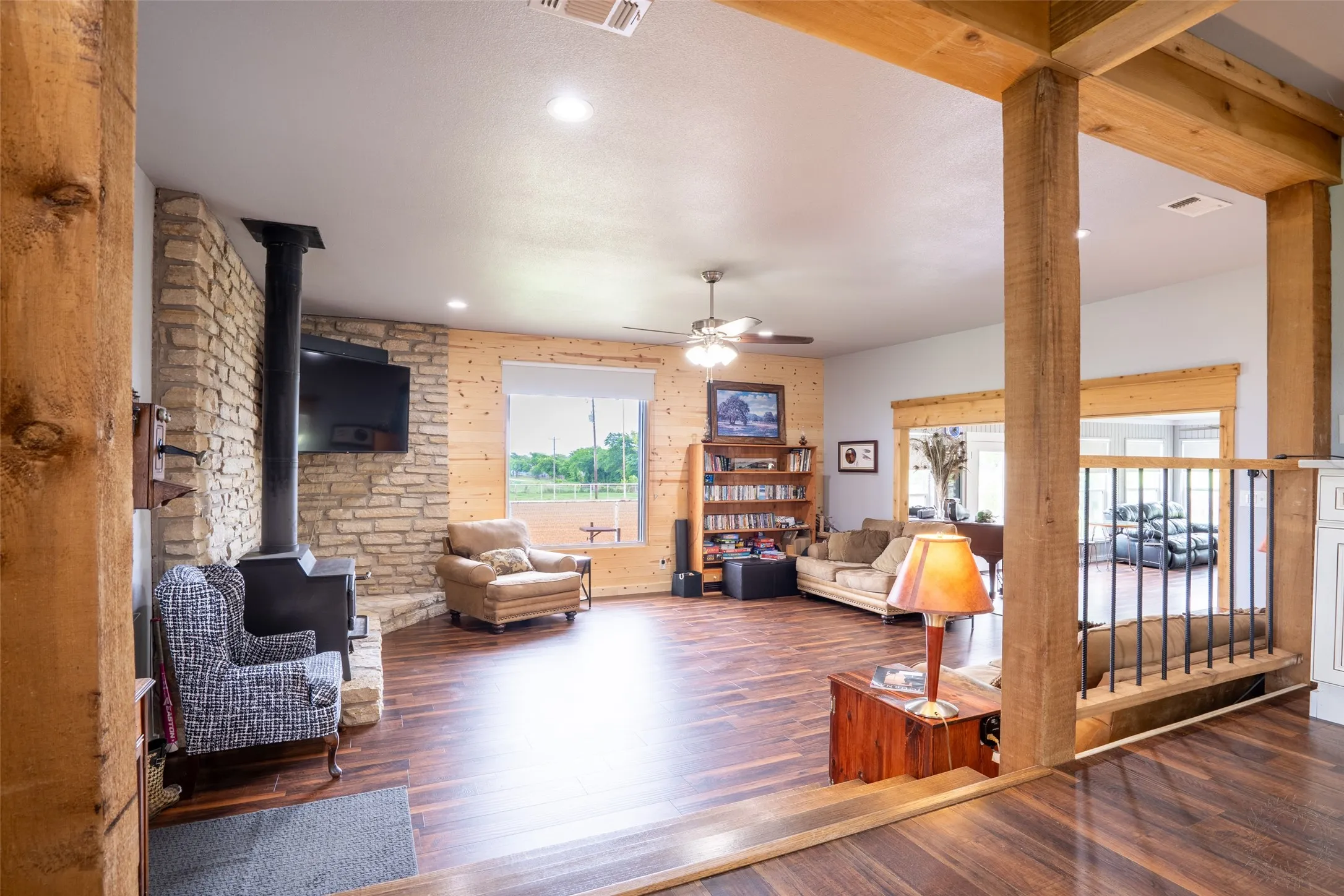 Spacious living area featuring wood-finish flooring, a stone fireplace with a built-in wood-burning stove, and exposed wood beam accents