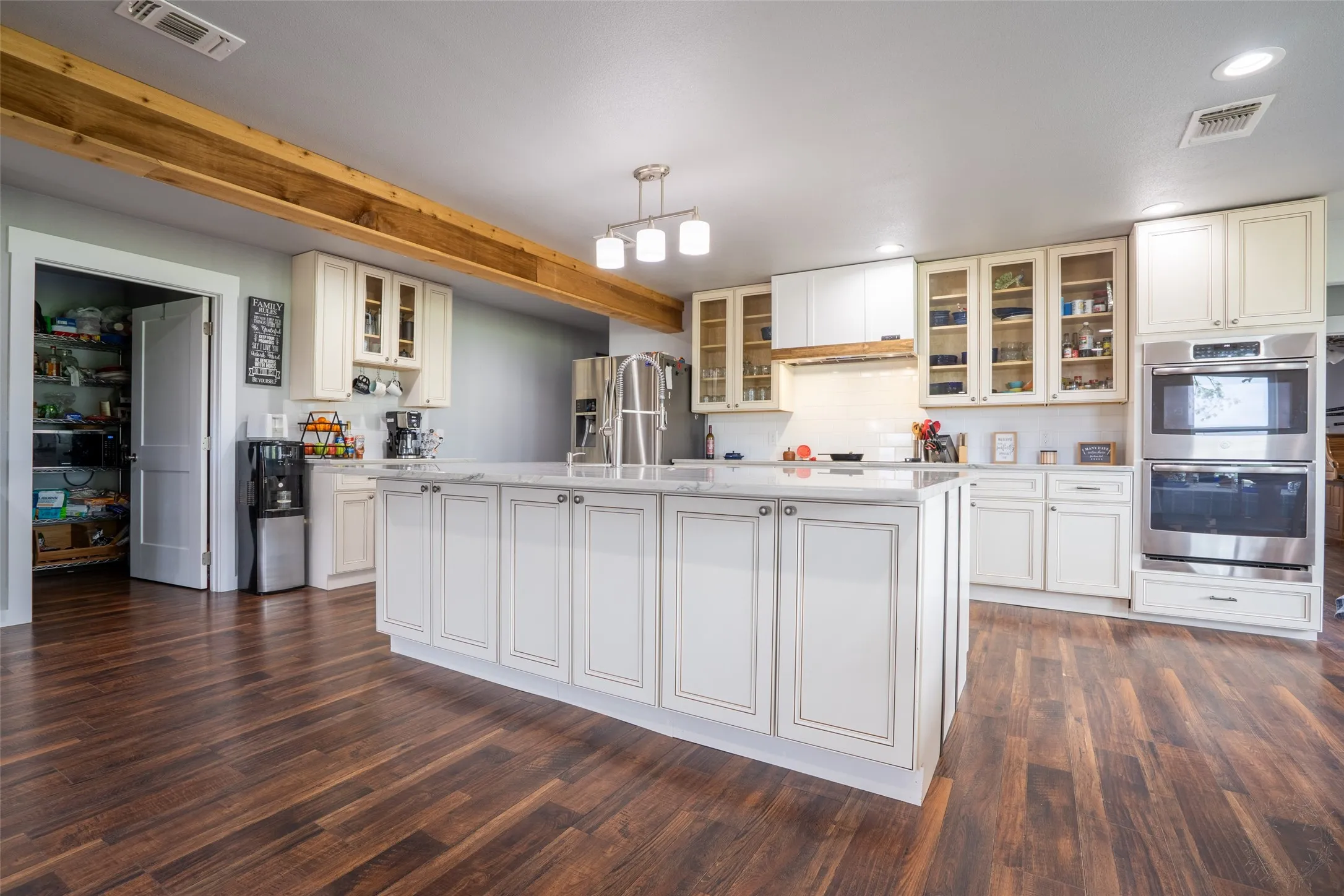 Kitchen featuring a central island with a light-toned countertop, wood-finish flooring, exposed ceiling beam, stainless steel double wall oven, and white cabinetry
