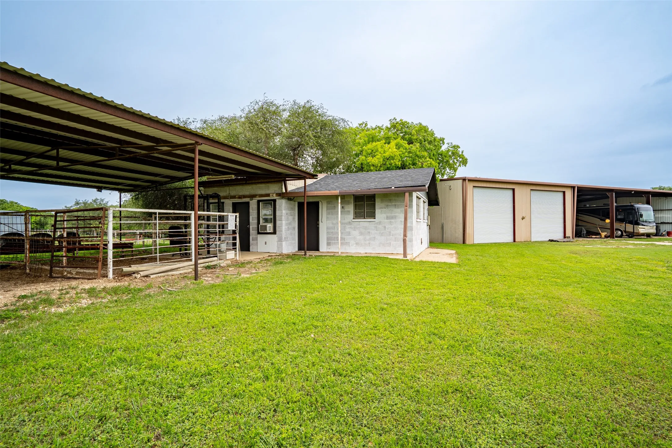 Expansive green space featuring multiple outbuildings including a covered metal structure, a block construction building with a window-mounted air conditioning unit, and a large metal garage building with two roll-up doors and an attached open-sided covered parking area