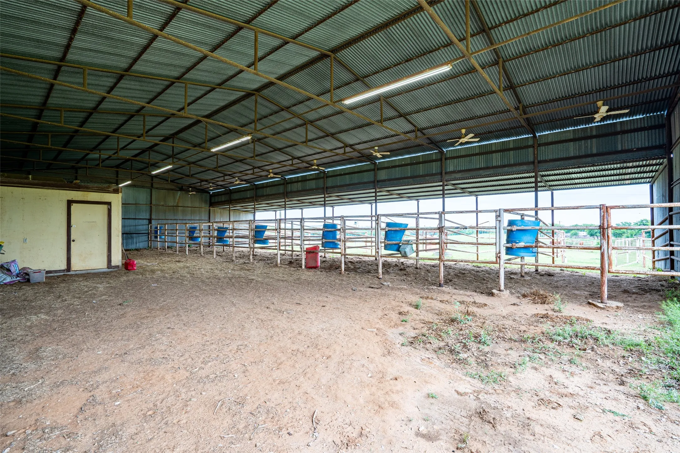 Metal-roofed barn structure featuring an open-air design, interior fencing, multiple fluorescent light fixtures, and ceiling fans