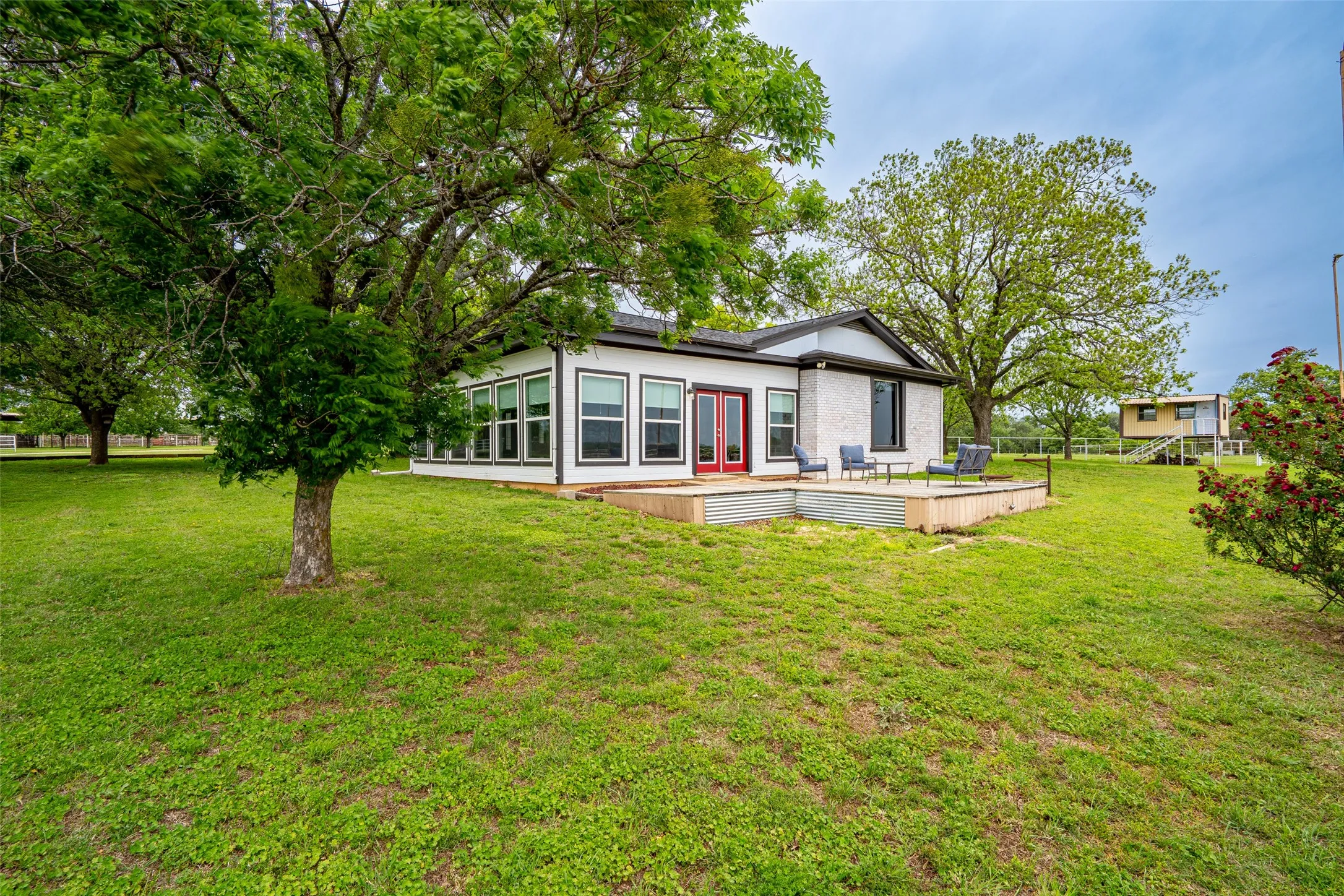 White brick exterior featuring a sunroom with multiple windows, a red double-door entry, a wooden patio deck, and mature trees in a grassy setting