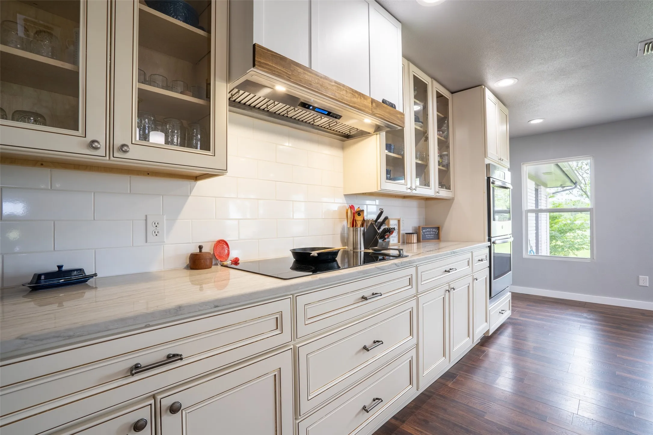 Kitchen featuring white cabinetry with glass-front uppers, light-toned countertops, a subway tile backsplash, and a built-in cooktop with a wood-accented range hood