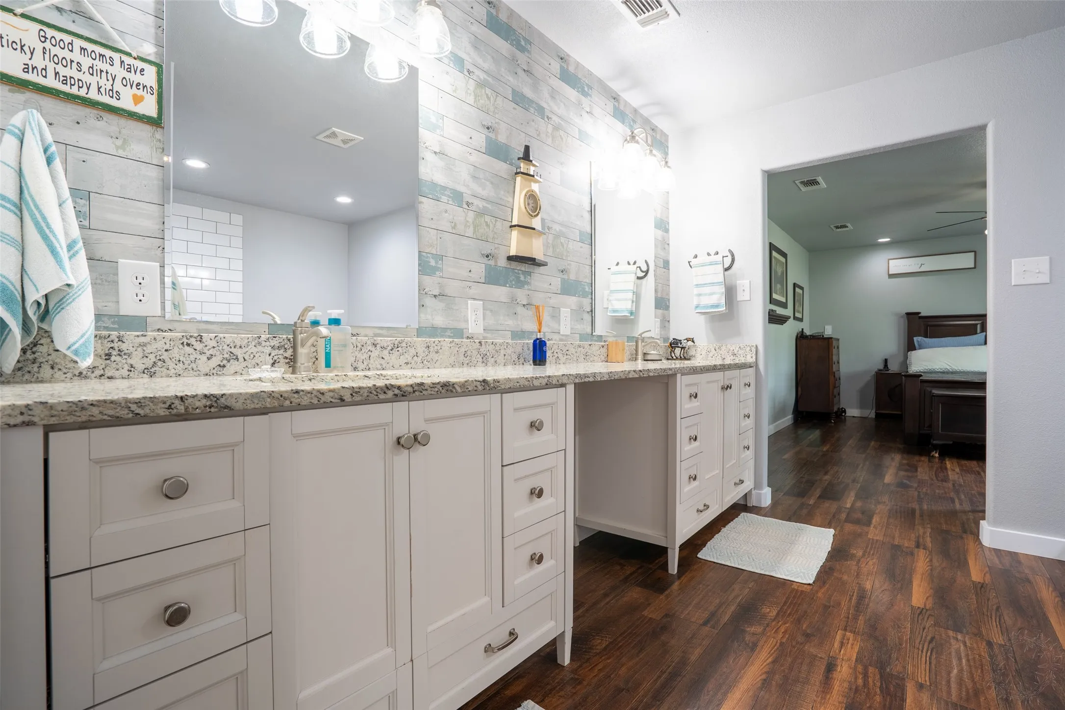 Dual vanity with granite countertops, light wood-finish shiplap accent wall, white cabinetry with silver hardware, and wood-finish flooring