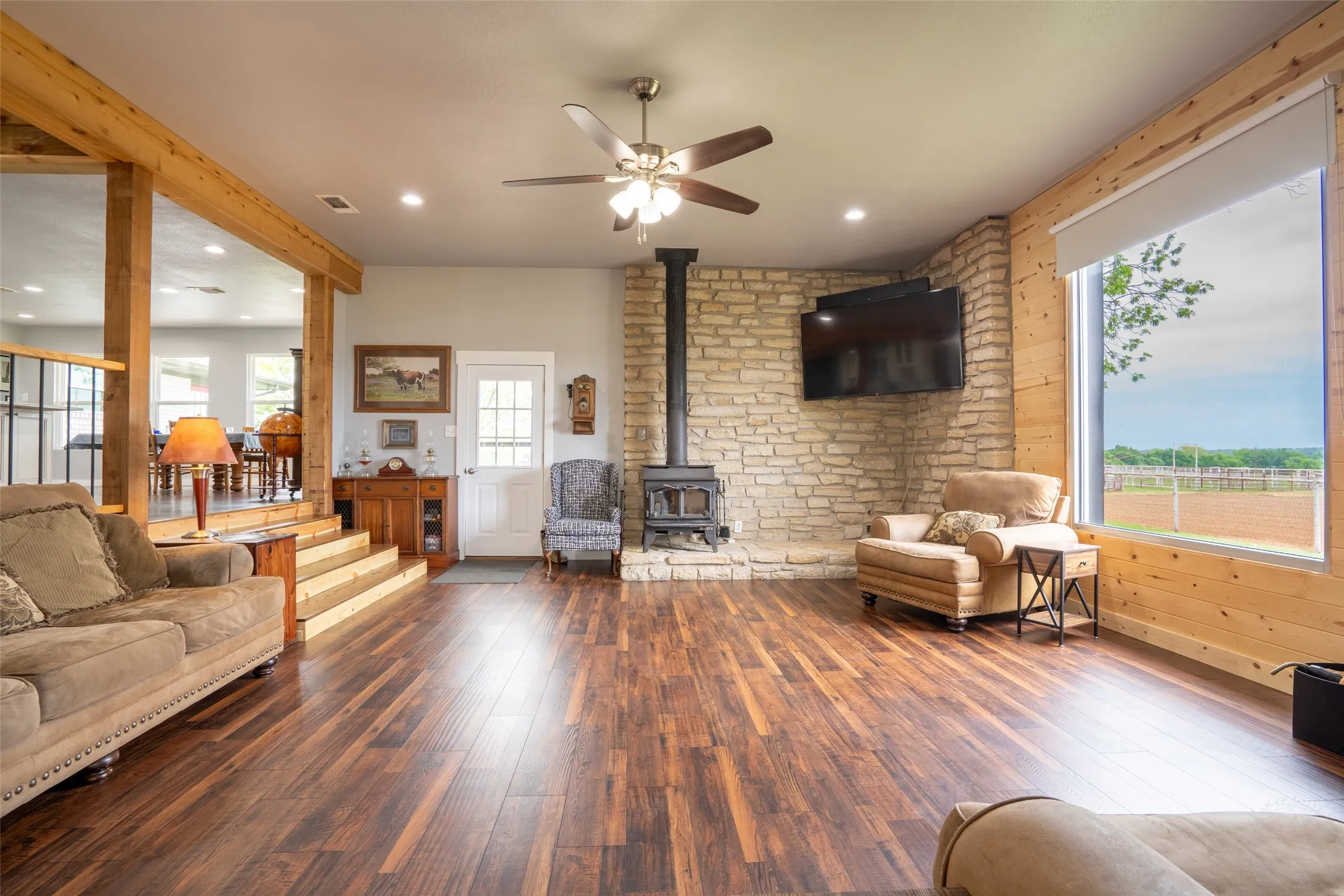 Spacious living area featuring rich wood-finish flooring, a stone fireplace with a wood-burning stove, and a ceiling fan with integrated lighting