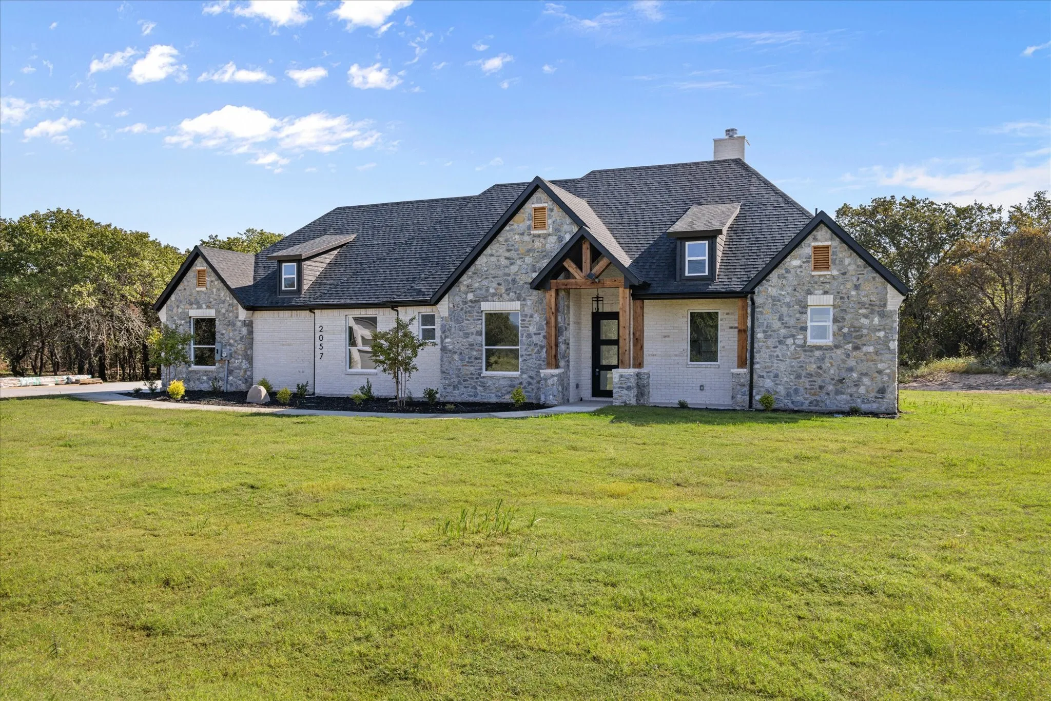 Grayish light-colored Stone and brick exterior with dark shingle roof and dormer windows