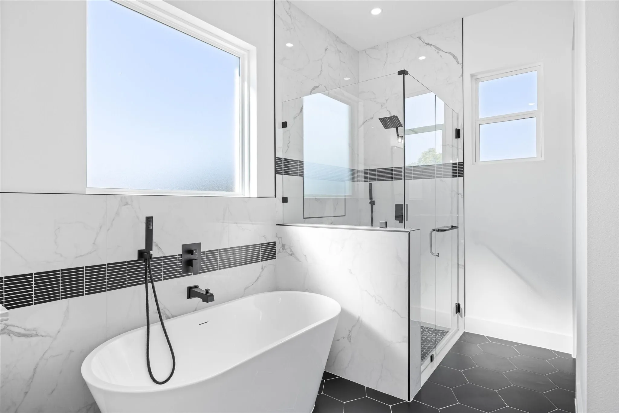 Freestanding soaking tub with matte black fixtures, marble-look tile walls with a dark accent band, frameless glass shower enclosure, and hexagonal floor tiles