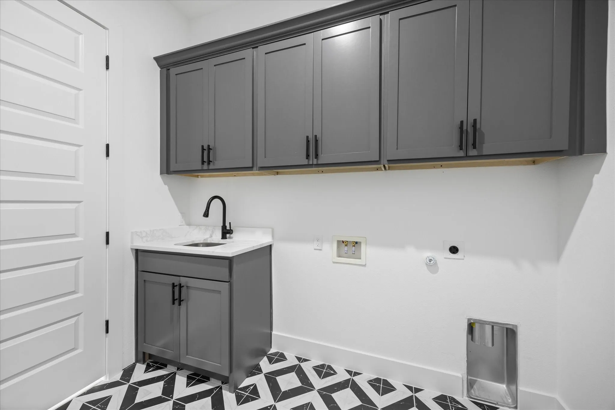 Laundry room featuring a white paneled door, dark gray shaker-style cabinetry, a white countertop with integrated sink, a matte black faucet, and patterned floor tiling