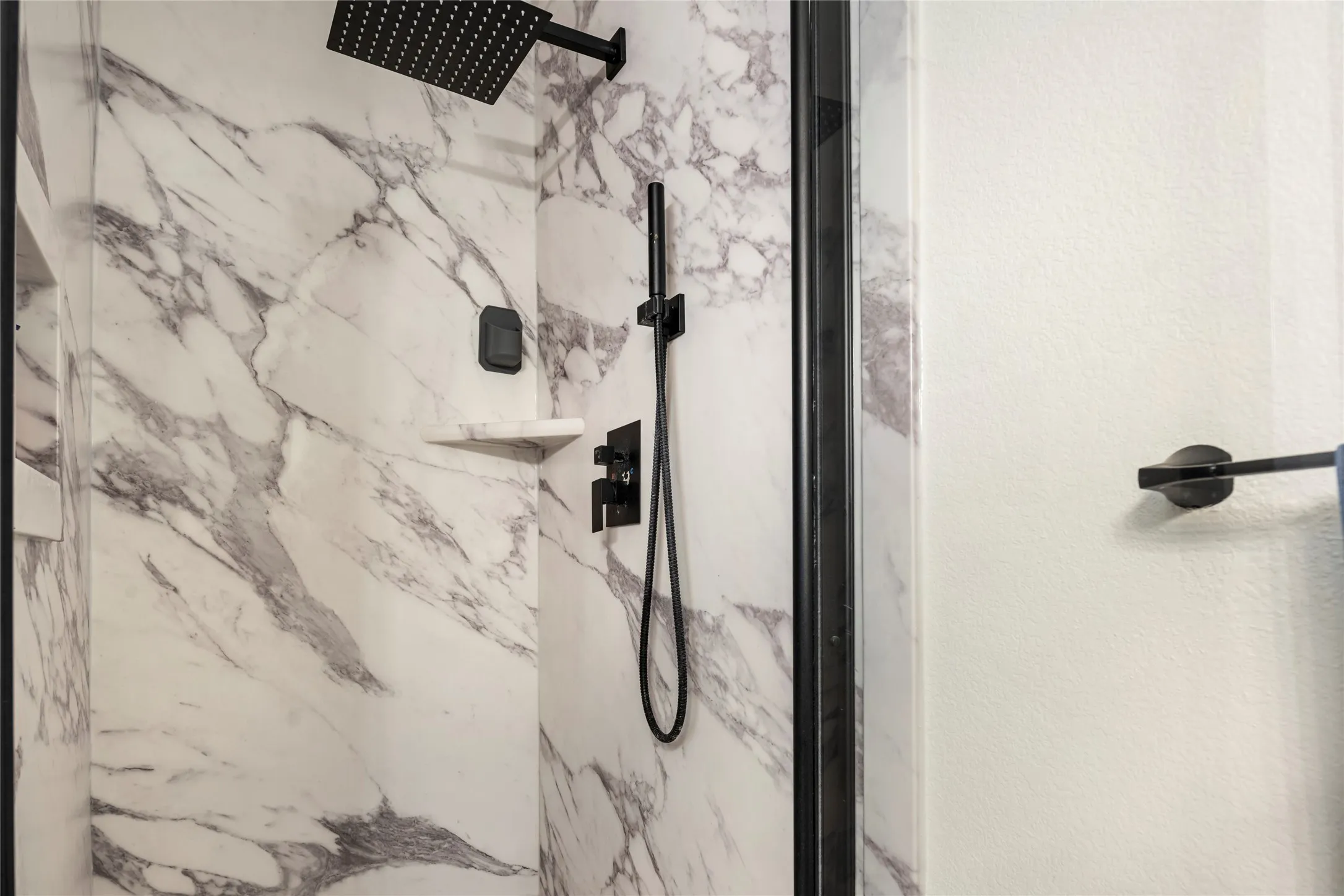 Shower enclosure featuring marble-patterned paneling, a rain showerhead, a handheld shower fixture, and a built-in corner shelf, all with matte black finishes