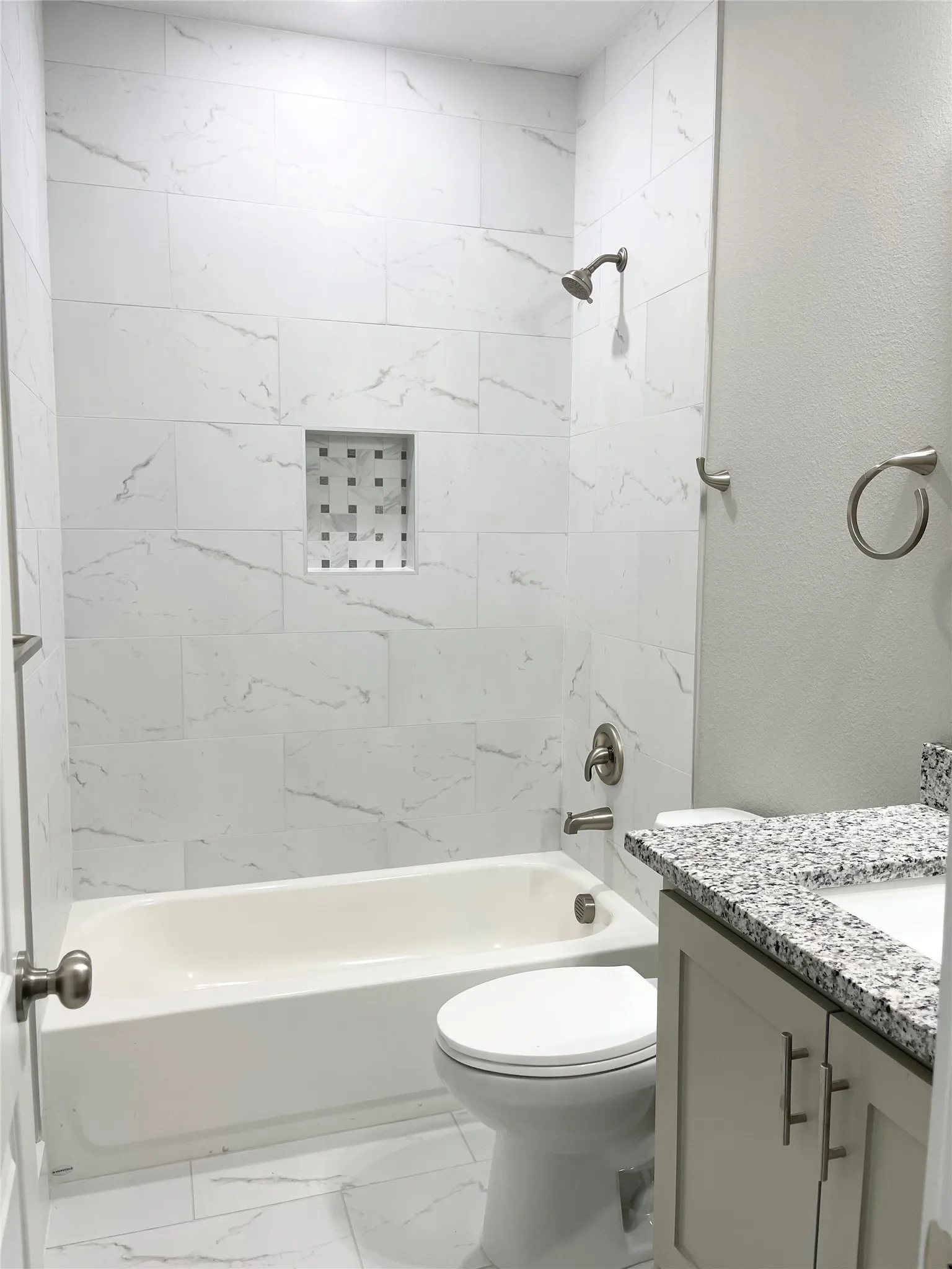 Bathroom featuring a white tub, white tiled shower surround with a mosaic-tiled niche, and a vanity with a granite countertop