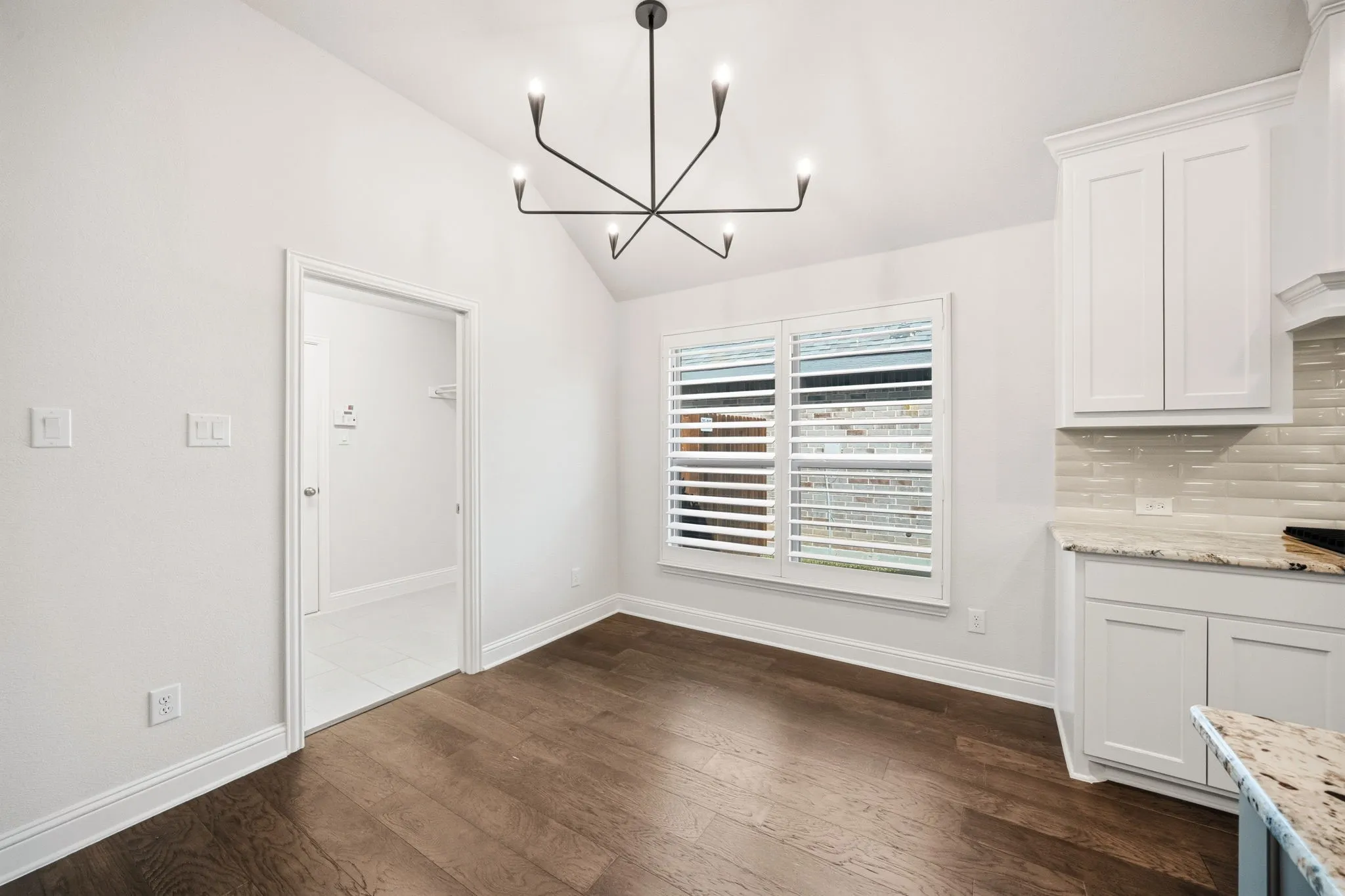 Vaulted ceiling with modern chandelier, wood-finish flooring, white plantation shutters, white cabinetry, and a light-toned backsplash