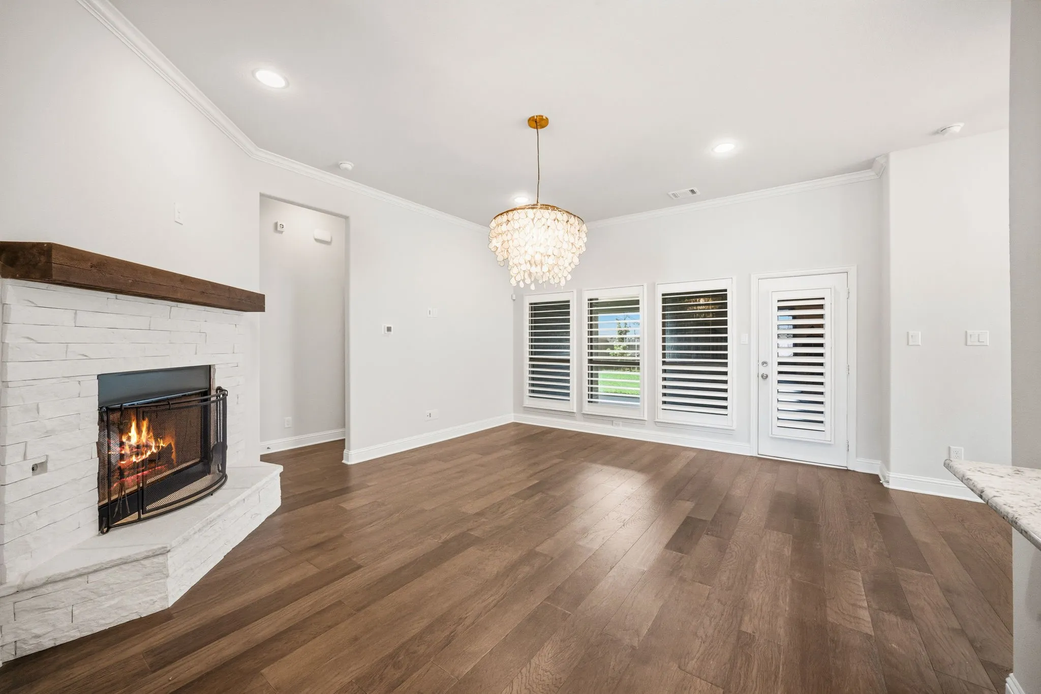 Spacious interior room featuring wood-finish flooring, a white stacked stone fireplace with a wood mantel, and multiple windows with plantation shutters