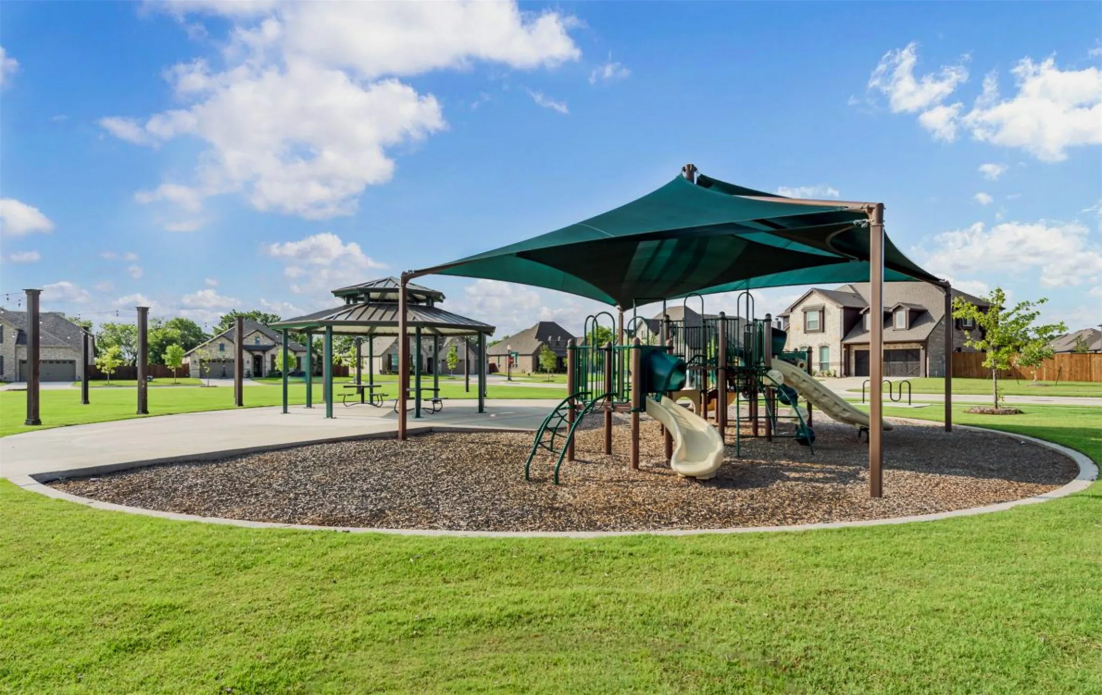 Community playground featuring a multi-level play structure with slides, a shaded canopy, a separate gazebo with picnic tables, a concrete pathway, and surrounding green space