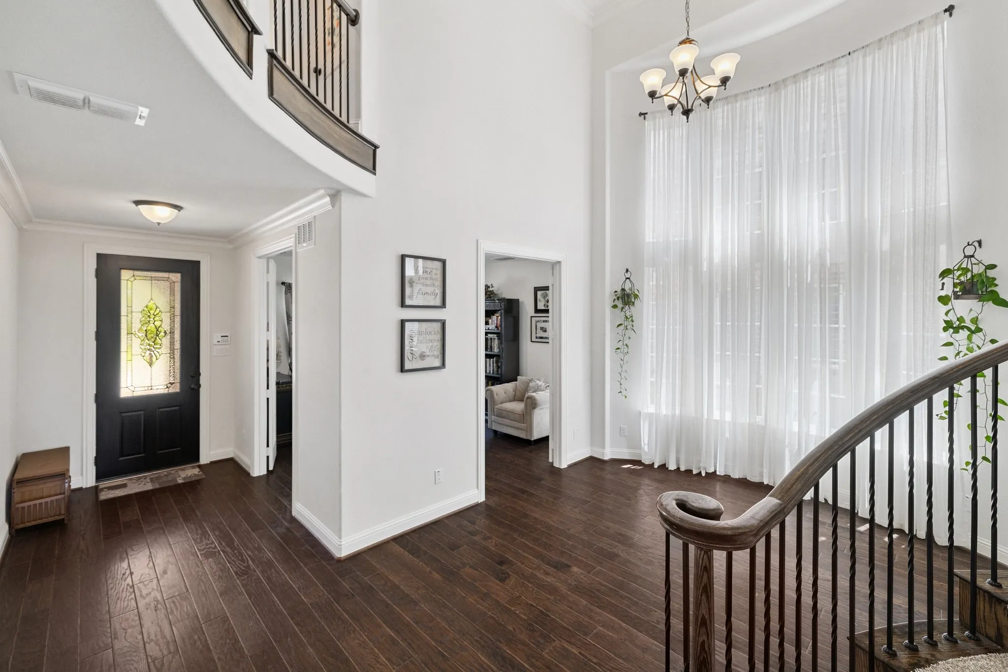 Foyer featuring a dark wood-finish door with leaded glass, dark wood-finish flooring, a two-story ceiling, a grand staircase with wrought iron balusters, and expansive windows with sheer curtains