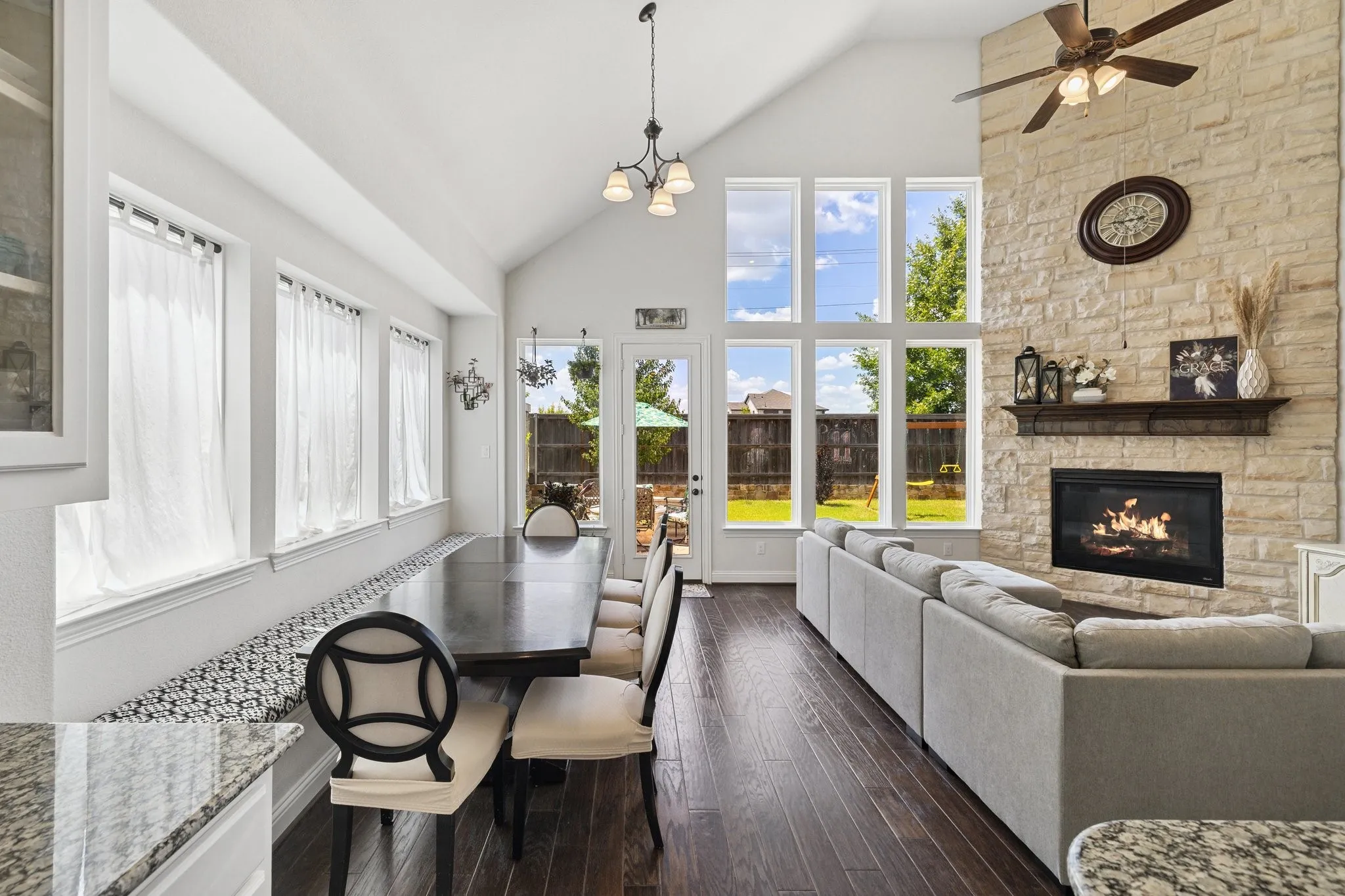 Vaulted ceiling open-concept room featuring wood-finish flooring, a stone-clad fireplace with a dark wood mantel, and multiple large windows overlooking the backyard
