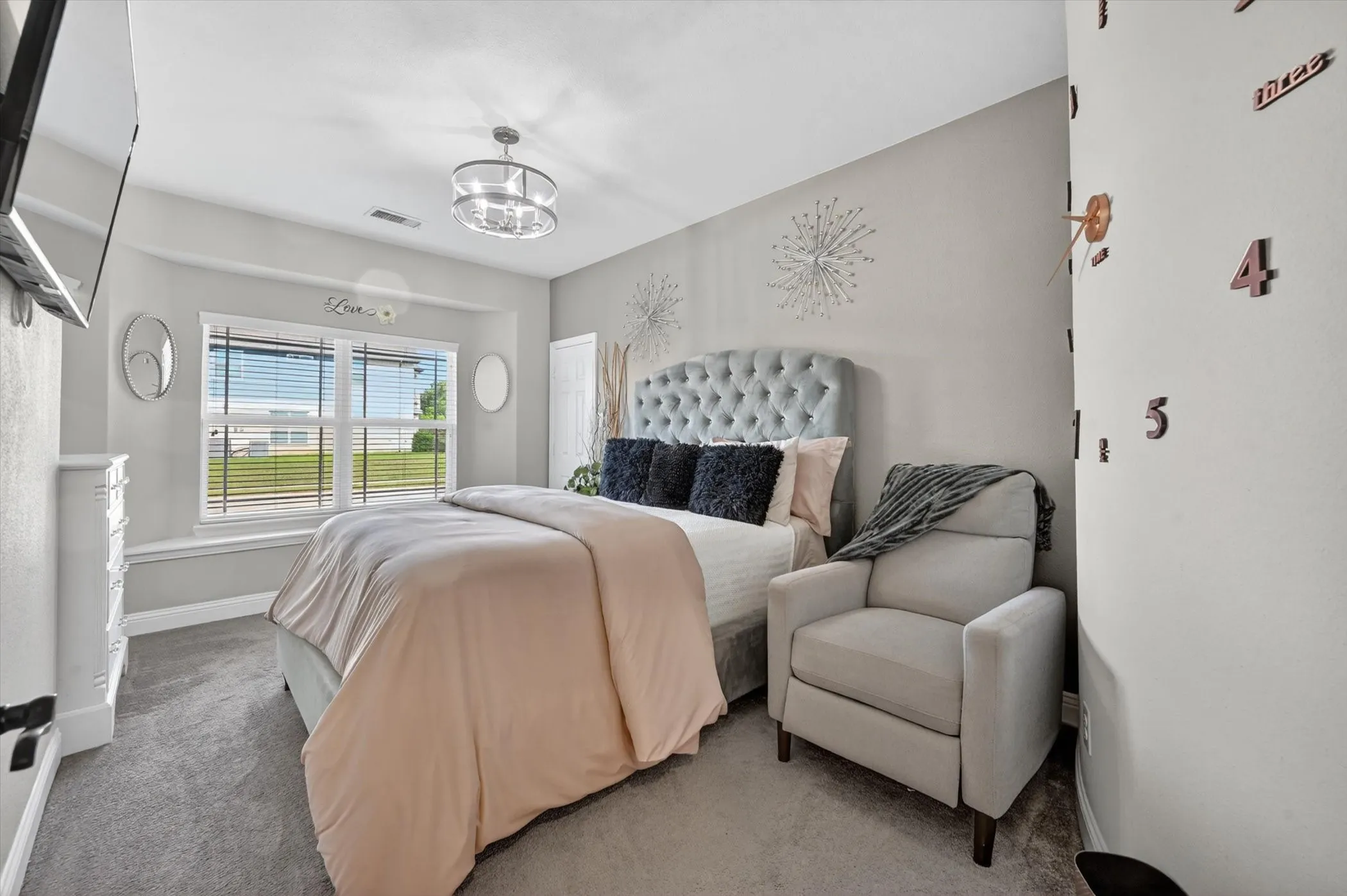 Carpeted bedroom featuring a large window with horizontal blinds, a tufted upholstered headboard, and a contemporary ceiling light fixture