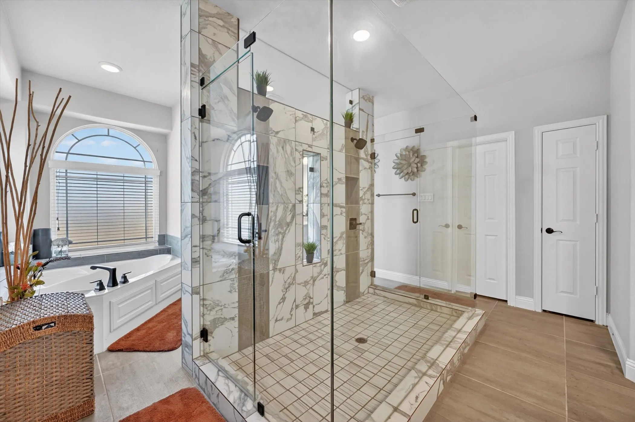 Primary Bath Spacious bathroom featuring a glass-enclosed shower with marble-look tile, a built-in soaking tub, an arched window, and wood-finish flooring