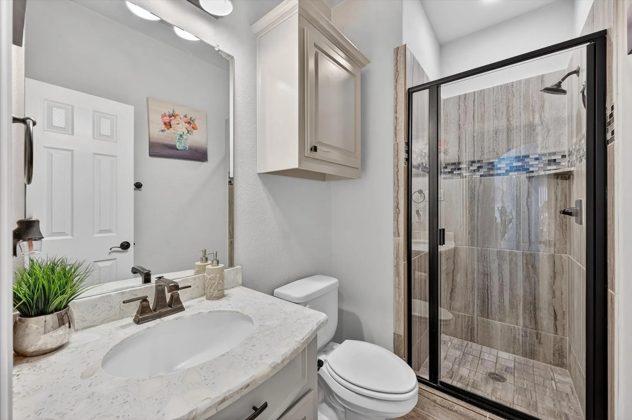 Light-filled bathroom featuring a single vanity with a white countertop and undermount sink, a framed mirror, and a glass-enclosed shower with vertical tile and a decorative mosaic band