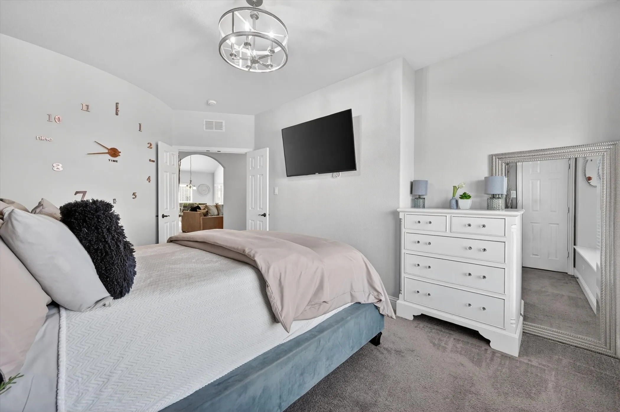 Spacious bedroom featuring light gray walls, plush gray carpeting, a contemporary ceiling light fixture, and arched doorways leading to another interior space