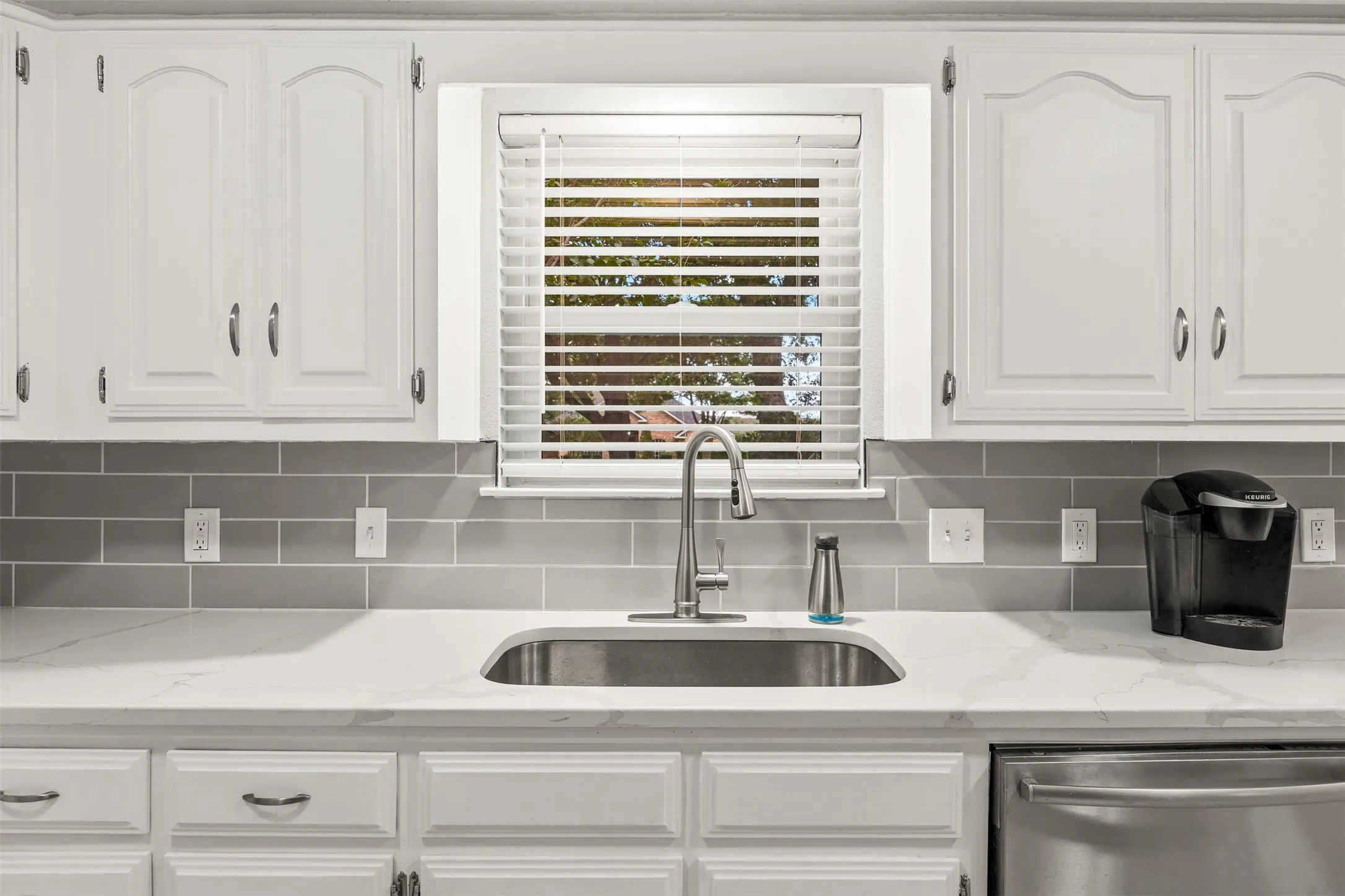 Kitchen featuring white cabinetry, a stainless steel sink, a brushed nickel faucet, light-toned countertops, and a gray subway tile backsplash