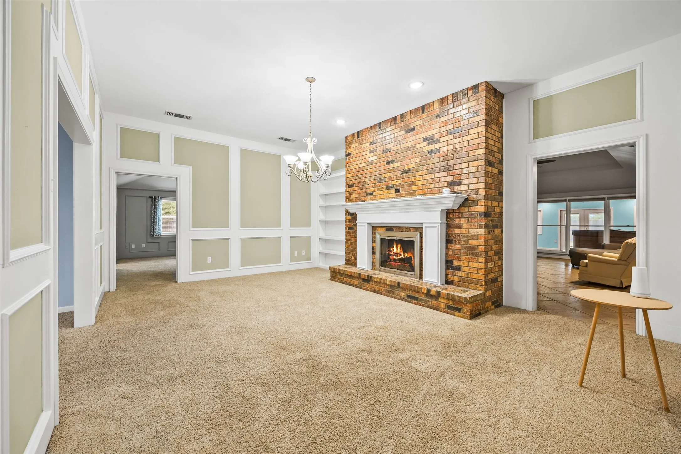 Expansive interior room featuring a brick fireplace with a white mantel, recessed lighting, a chandelier, wall paneling, and built-in shelving