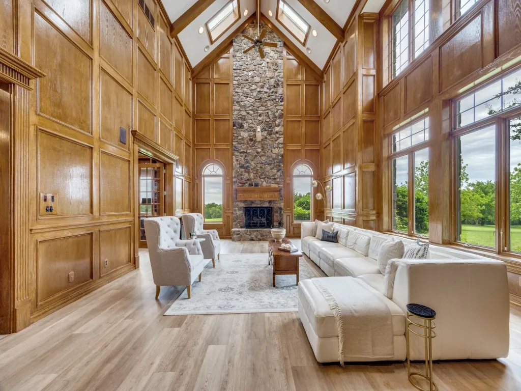 Soaring vaulted ceilings with skylights, a floor-to-ceiling natural stone fireplace, extensive wood-paneled walls, large windows, and light wood-finish flooring