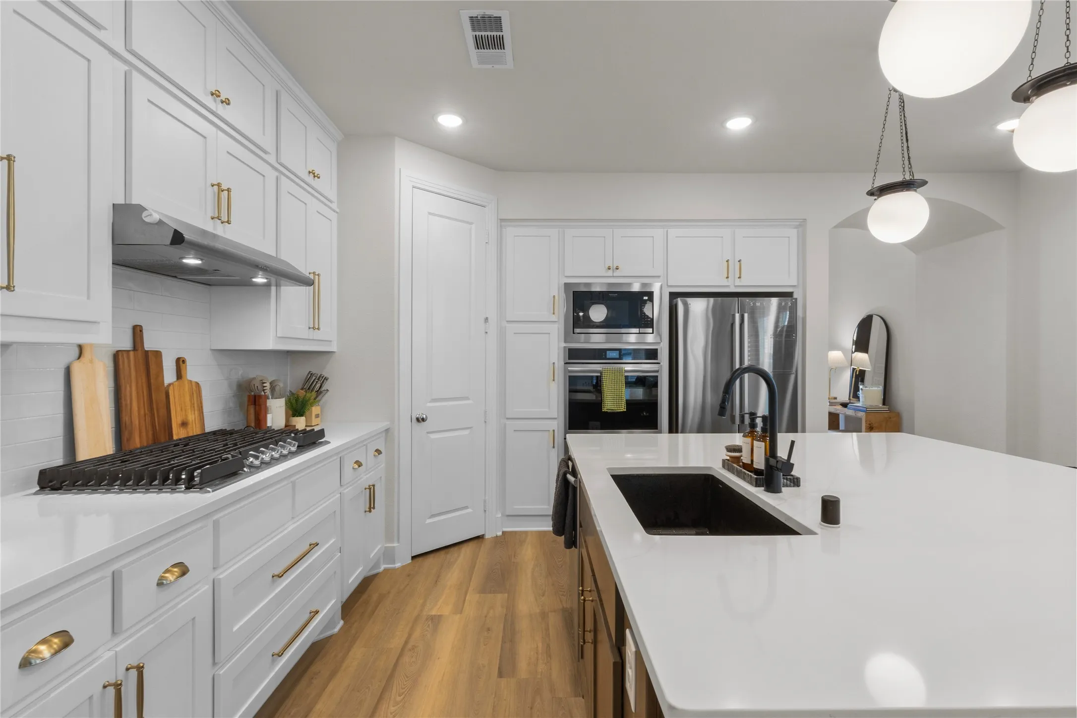 Modern kitchen featuring white cabinetry with gold-tone hardware, wood-finish flooring, a built-in stainless steel oven and microwave, and a large island with a black sink and matte black faucet