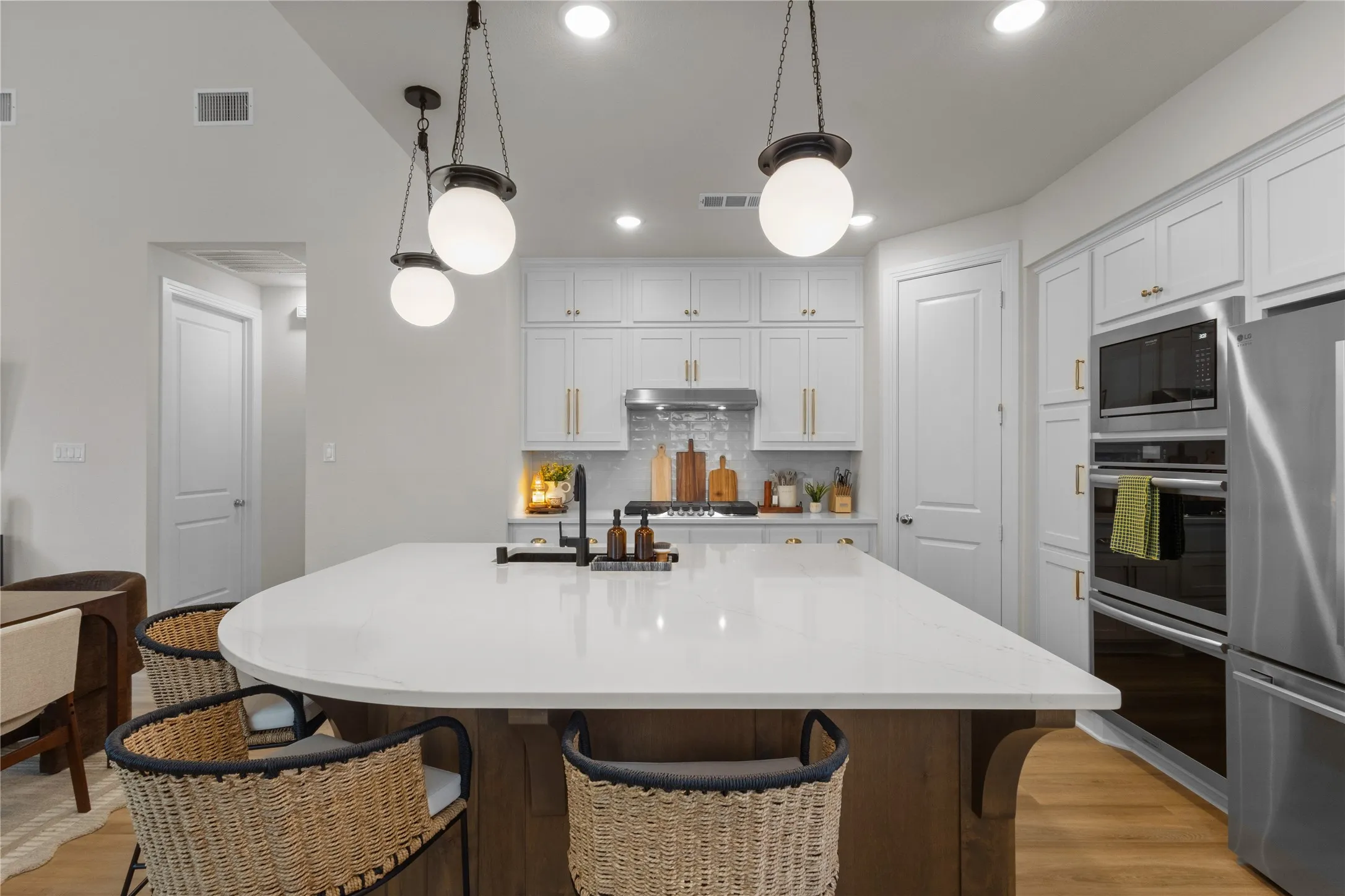 Spacious kitchen featuring a large central island with a white countertop and wood-finish base, dual pendant lighting, recessed ceiling lights, white cabinetry with gold-tone hardware, and integrated stainless steel appliances