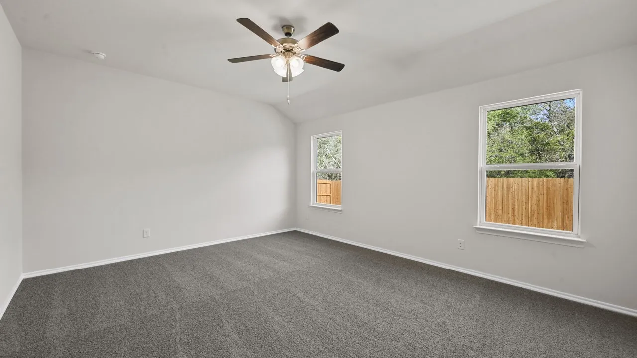 Spacious room featuring vaulted ceilings, a ceiling fan with light fixture, gray carpeting, and two windows overlooking a wooden fence and mature trees