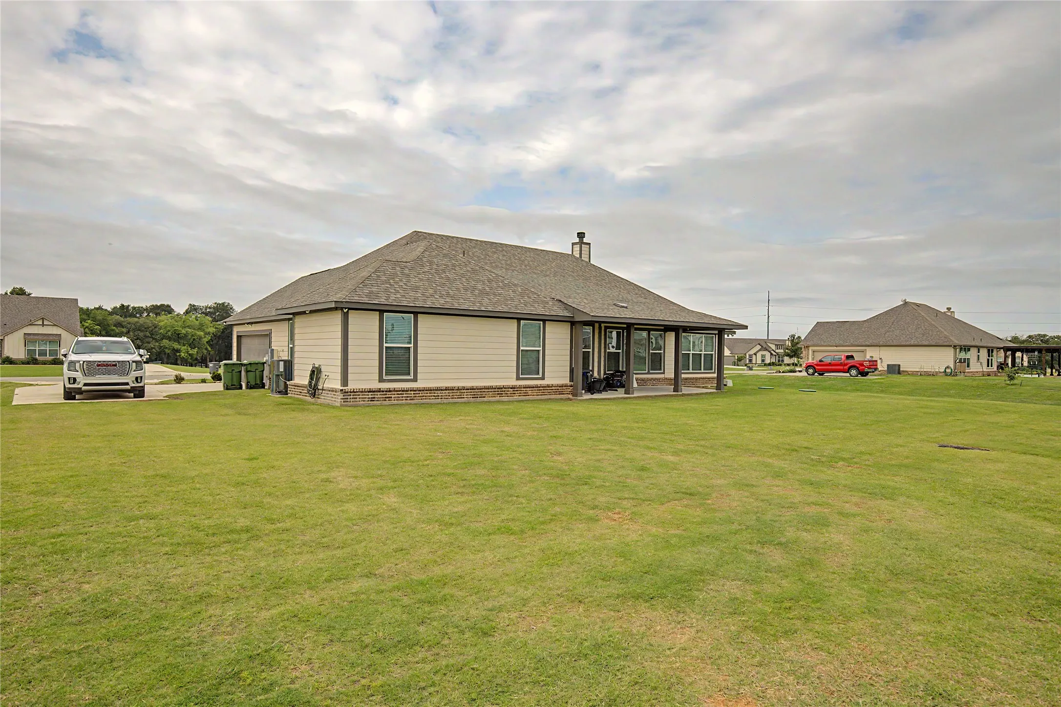 Expansive green lawn surrounds the property, featuring a covered patio with dark posts, multiple windows, and a side-entry garage